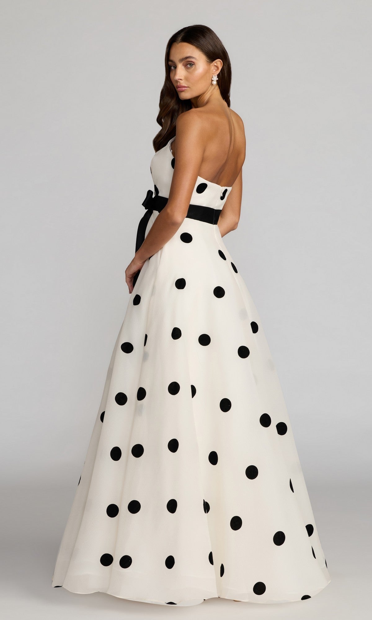 A woman wearing the Long Formal Dress: Mac Duggal 27834— a sleeveless white gown with large black polka dots and a black waistband—stands, looking over her shoulder.